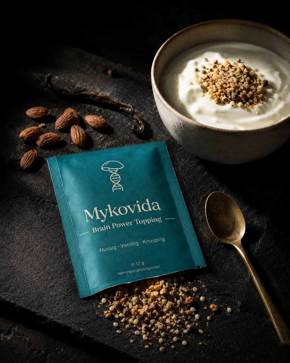 Petrolfarbenes Sachet Brain Power Topping neben einer Schale Joghurt mit nussigem Streusel, Mandeln und Vanilleschote auf dunklem Schiefer